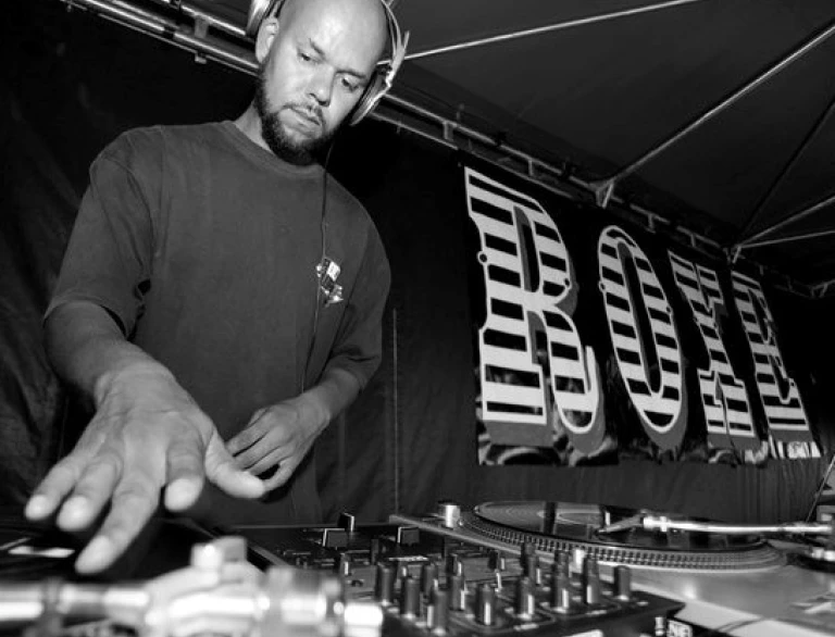 A DJ wearing headphones performs on a turntable setup under a tent, with a large "ROXE" banner in the background.
